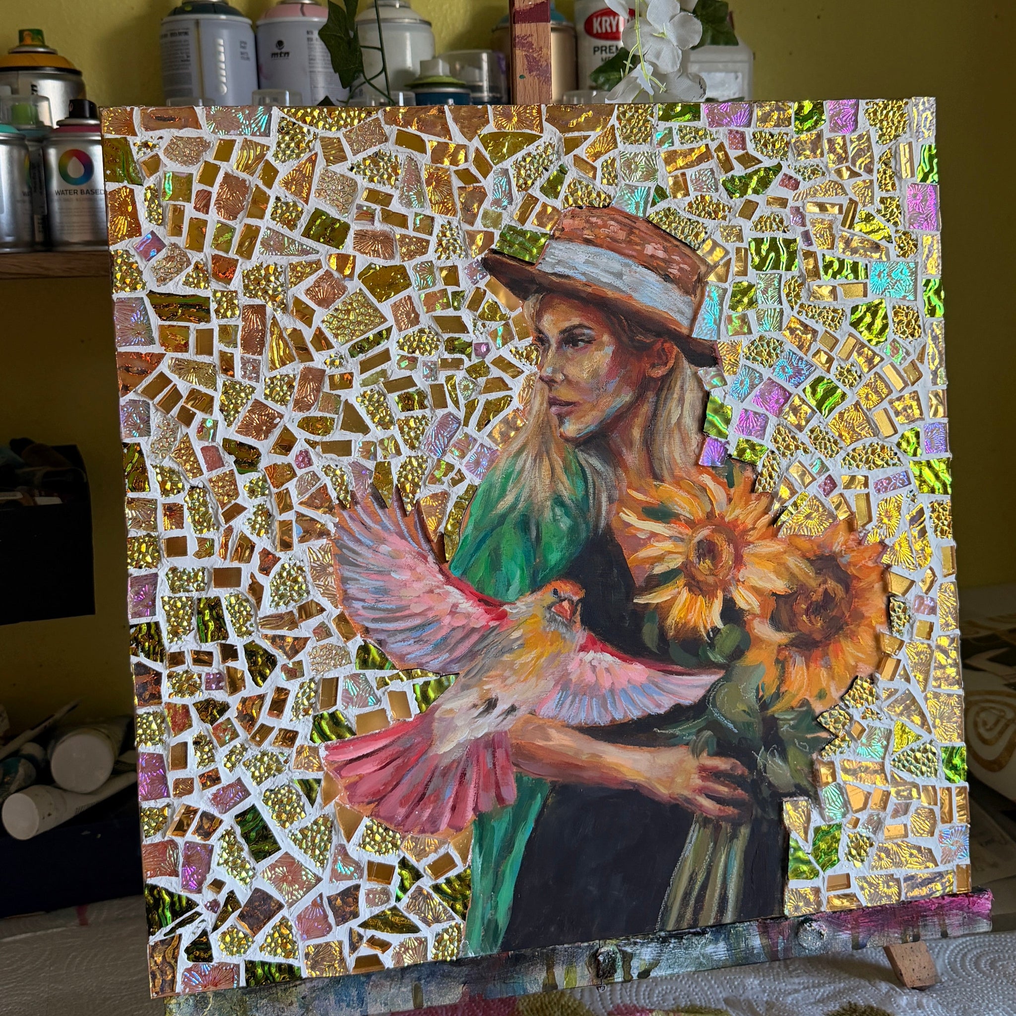 Mosaic artwork of a woman holding flowers on an easel with a yellow wall and plants in the background.