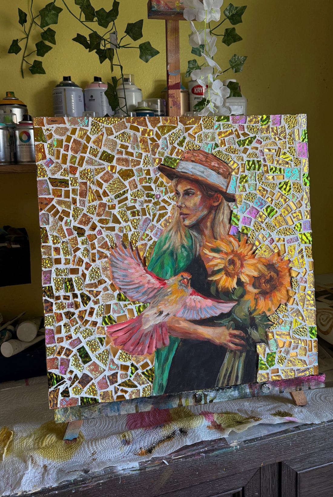 Mosaic artwork of a woman holding flowers on an easel with a yellow wall and plants in the background.