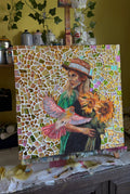 Mosaic artwork of a woman holding flowers on an easel with a yellow wall and plants in the background.