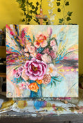 Colorful floral painting on an easel against a yellow wall with greenery.