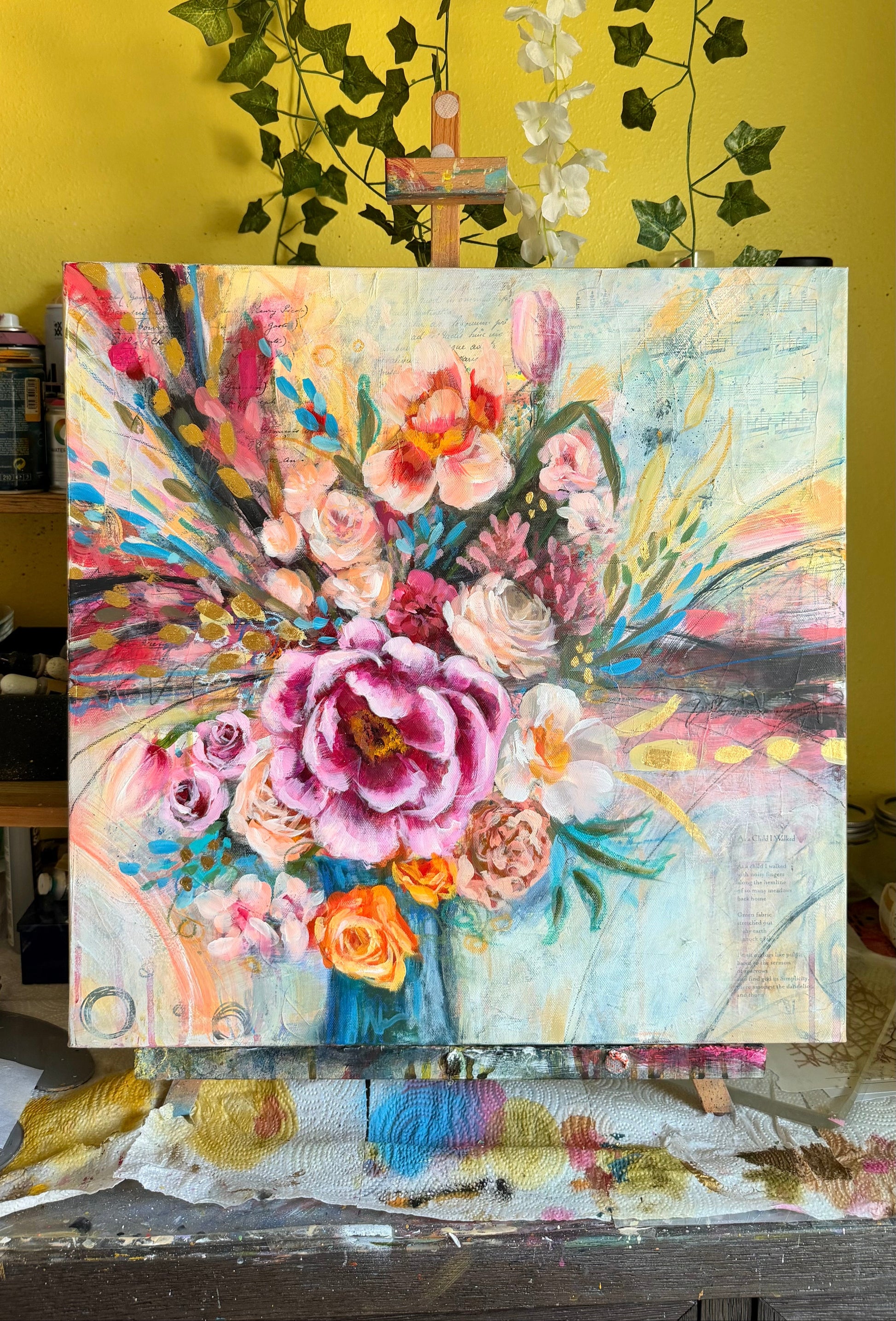 Colorful floral painting on an easel against a yellow wall with greenery.