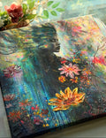 A colorful painted canvas featuring a dark silhouette portrait of a woman surrounded by vivid flowers and flowing blue-green textures, styled on a tabletop with flowers in the background.