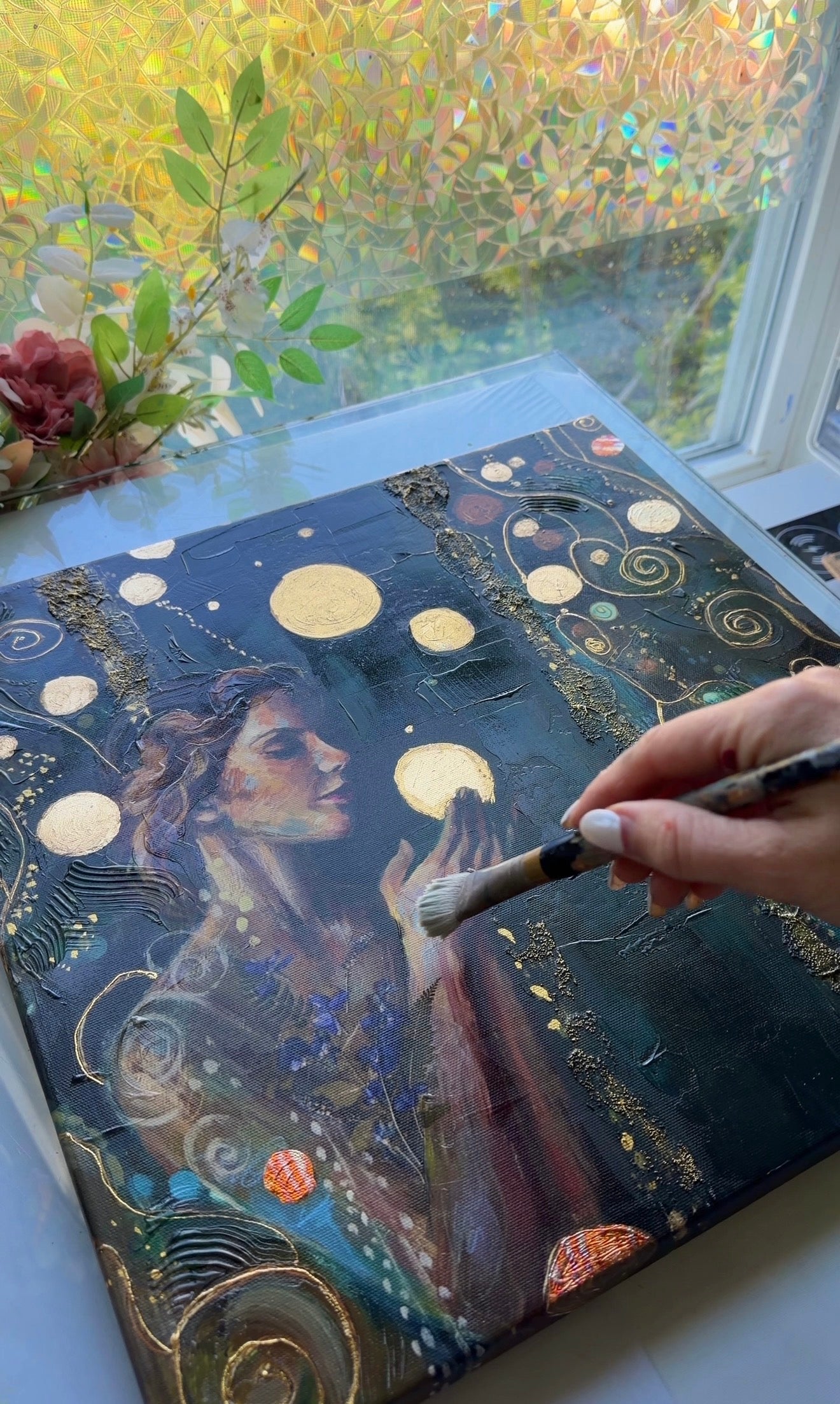 Artistic depiction of a woman with celestial elements on a canvas, held by a hand with a brush.