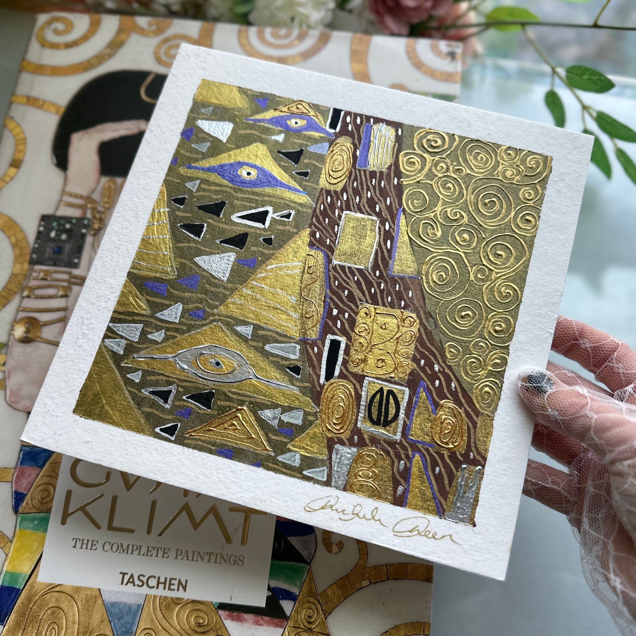 Artistic card with geometric design held by a gloved hand, with a book titled 'Klimt' in the background.