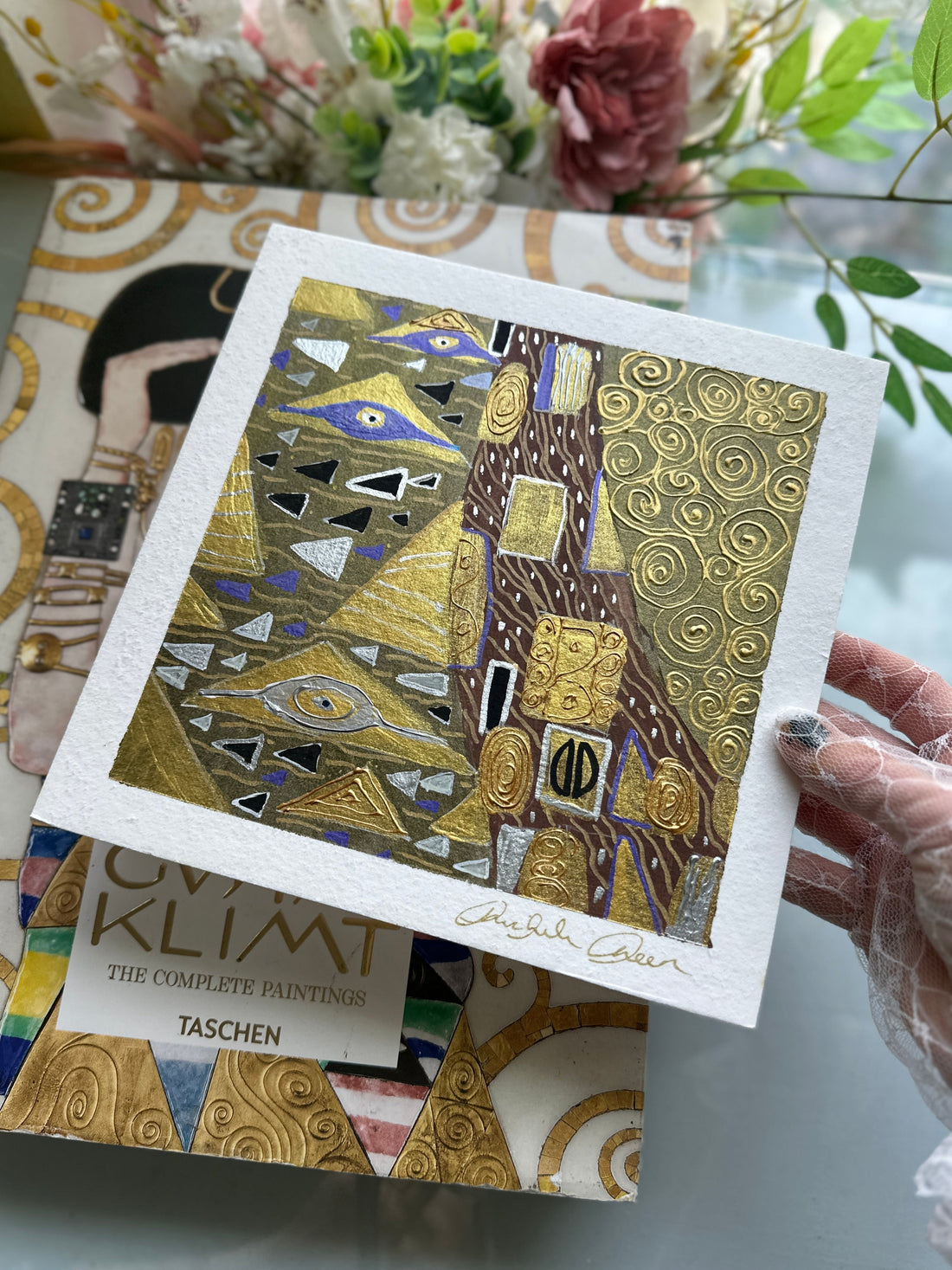 Artistic card with geometric design held by a gloved hand, with a book titled 'Klimt' in the background.