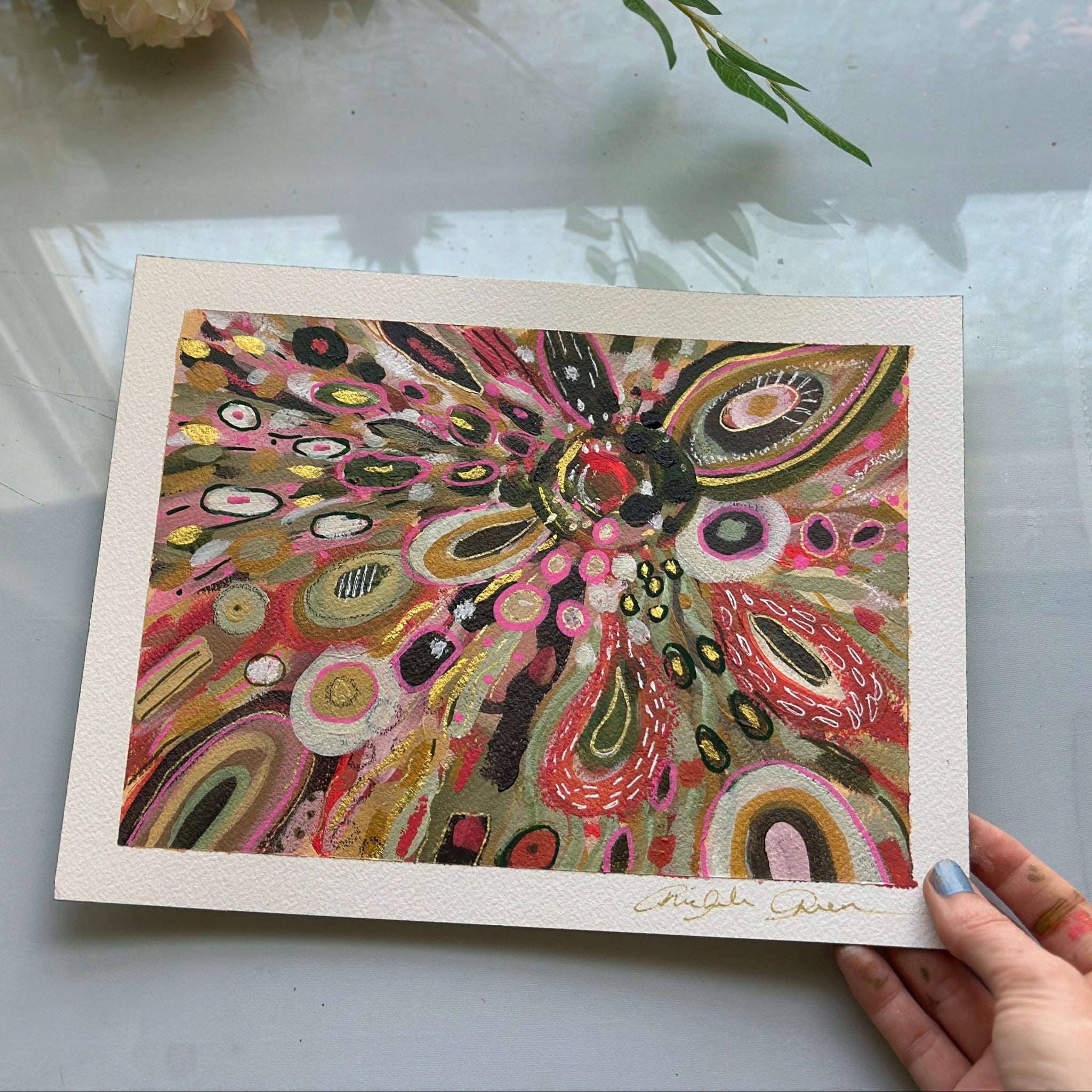Colorful abstract artwork held by a hand on a light surface with flowers in the background