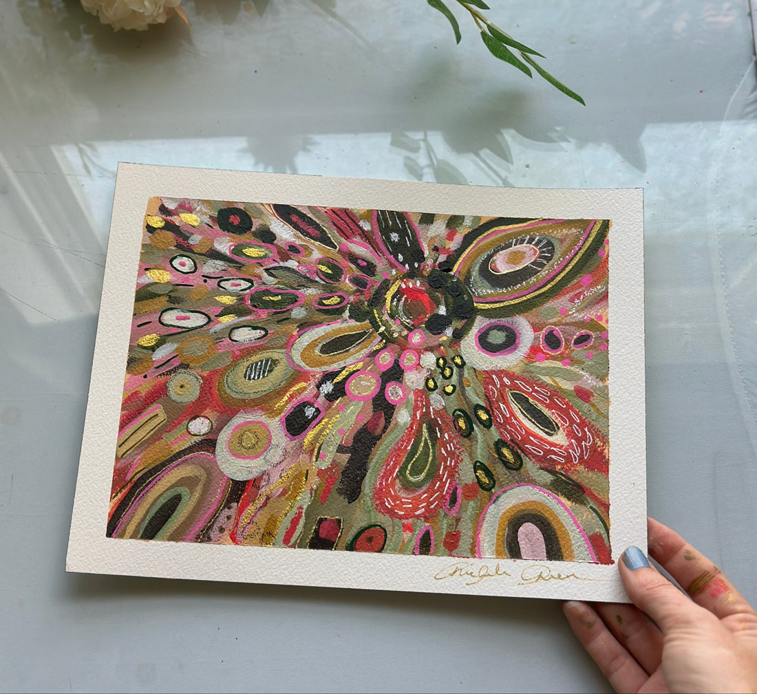 Colorful abstract artwork held by a hand on a light surface with flowers in the background