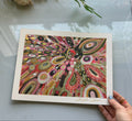 Colorful abstract artwork held by a hand on a light surface with flowers in the background