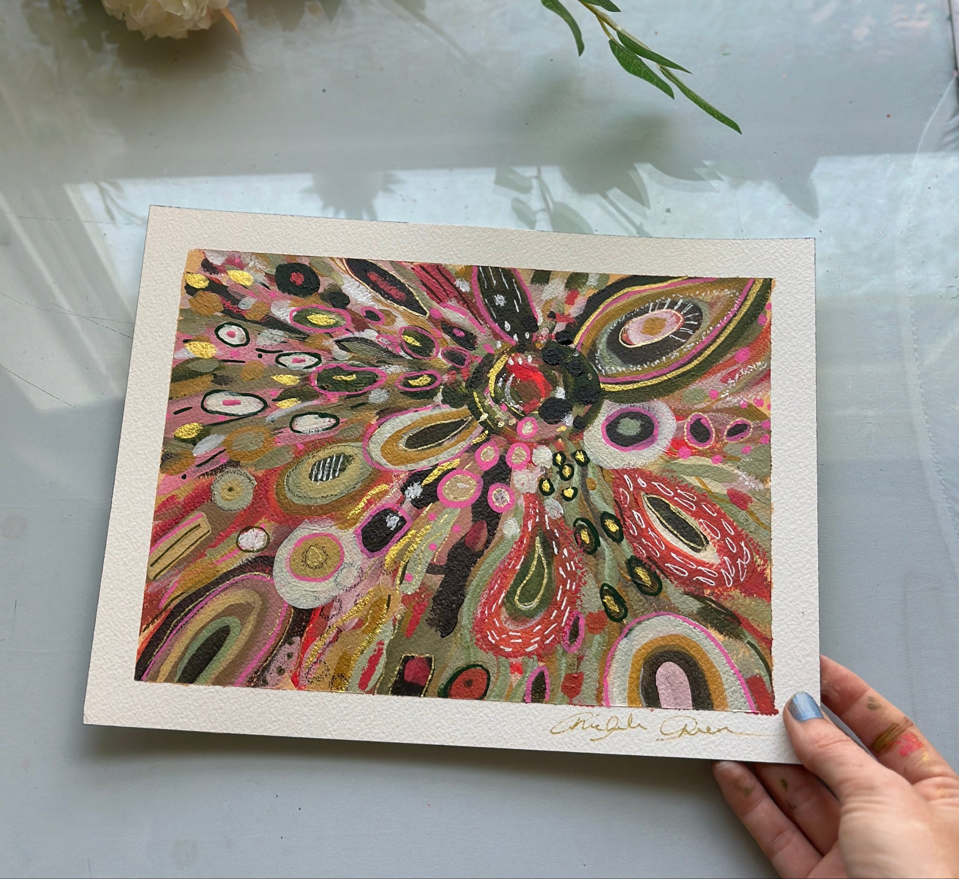 Colorful abstract artwork held by a hand on a light surface with flowers in the background