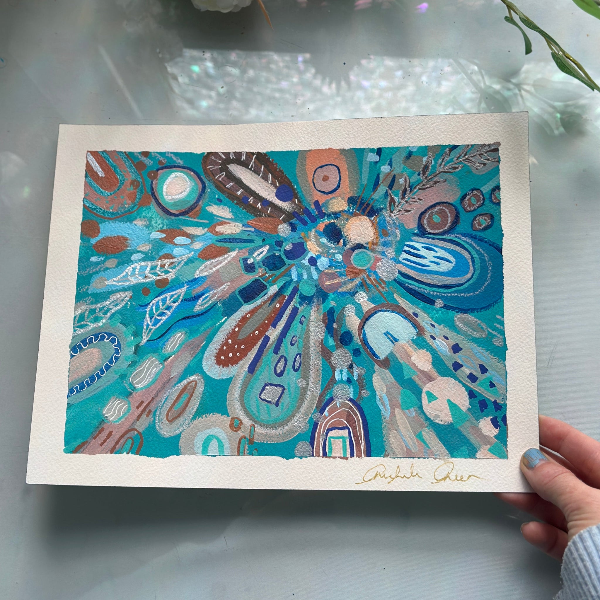 Colorful abstract artwork held by a hand on a reflective surface with flowers in the background