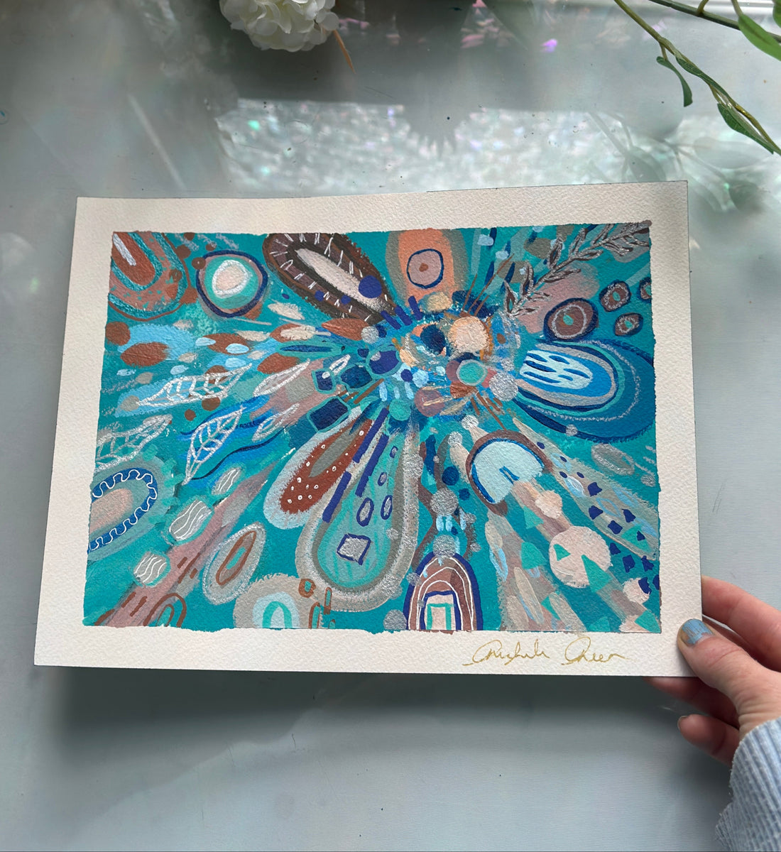 Colorful abstract artwork held by a hand on a reflective surface with flowers in the background