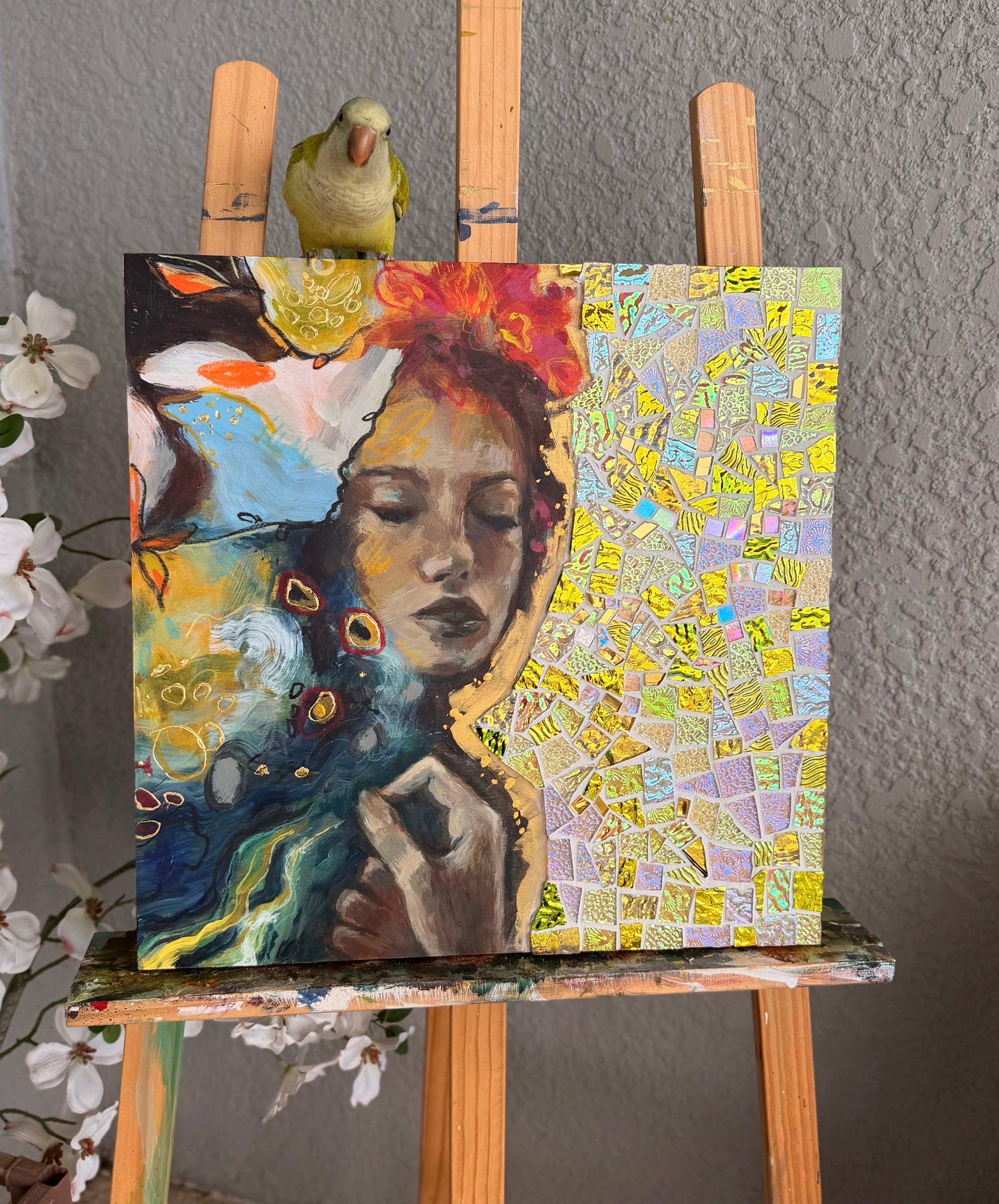 Colorful mosaic art on a wooden easel with a bird perched on top against a gray wall.