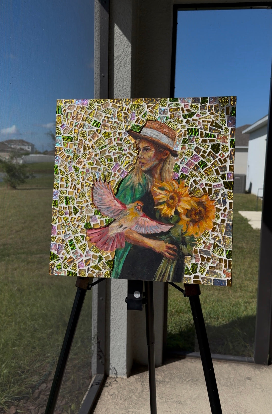 Mosaic artwork of a woman holding flowers on an easel with a yellow wall and plants in the background.